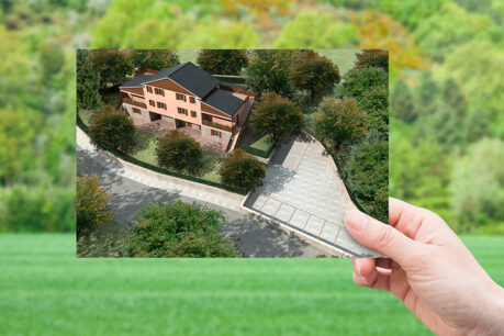 Hand holding up image of new home in front of an open area of land