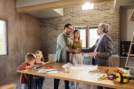 Man and woman couple shaking hands with lead builder as workers work on constructing home