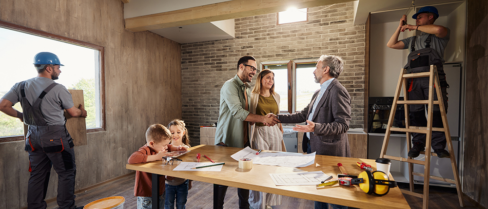 Man and woman couple shaking hands with lead builder as workers work on constructing home