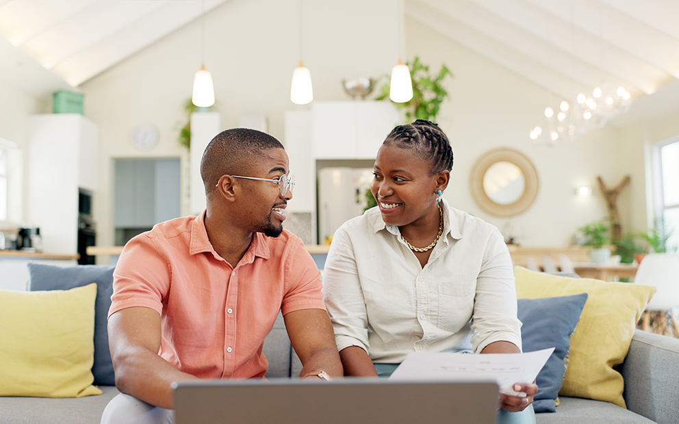 Man and woman coupling sitting on couch while smiling and discussing over laptop