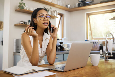 Woman talking on the phone while sitting in her kitchen with laptop on the table in front of her