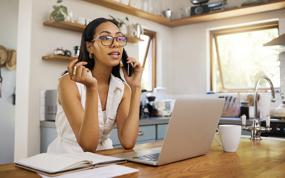 Woman talking on the phone while sitting in her kitchen with laptop on the table in front of her