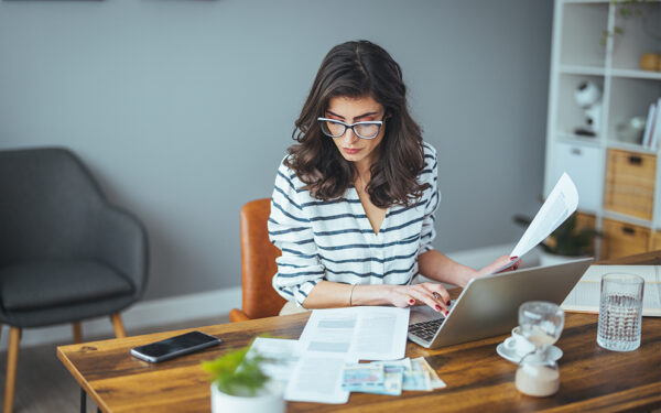 Woman sitting at home desk looking over laptop and documents